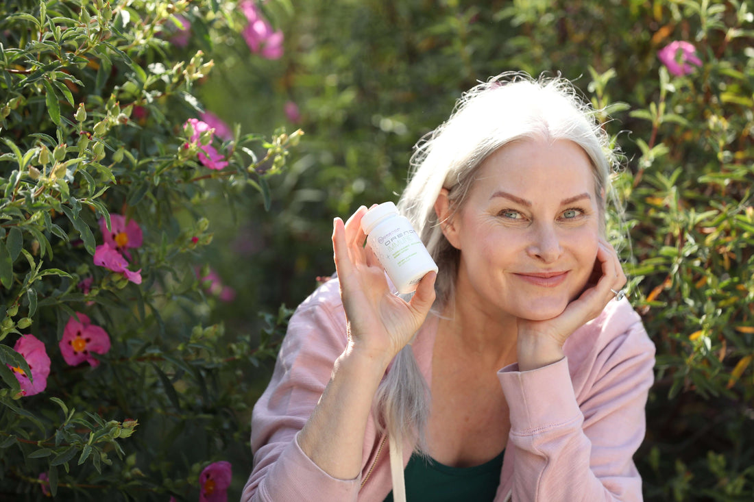 Wwoman smiling holding a bottle of Orenda Immune in front of hedge of flowering bush