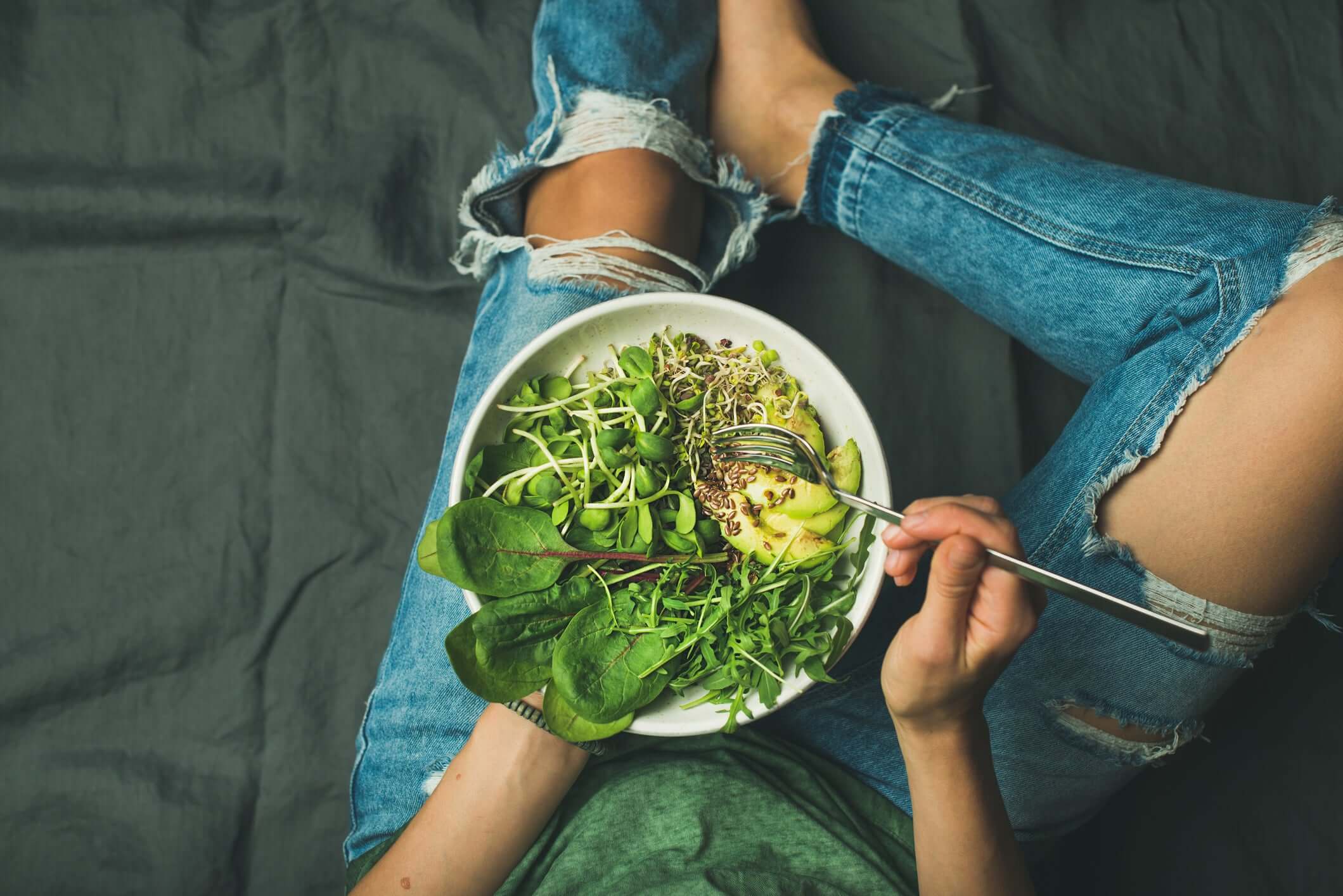 Girl in torn jeans sitting with a bowl of salad on her lap holding a fork